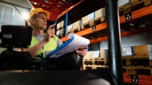 Trabajador revisando inventario en almacén logístico con estanterías y palets en un centro de logística empresarial.