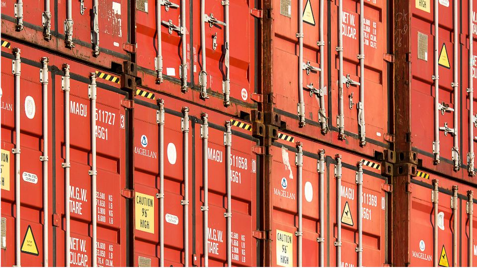 Contenedores de mercancías rojos apilados en una terminal logística, utilizados para ilustrar los factores que influyen en el precio del transporte de mercancías por carretera.